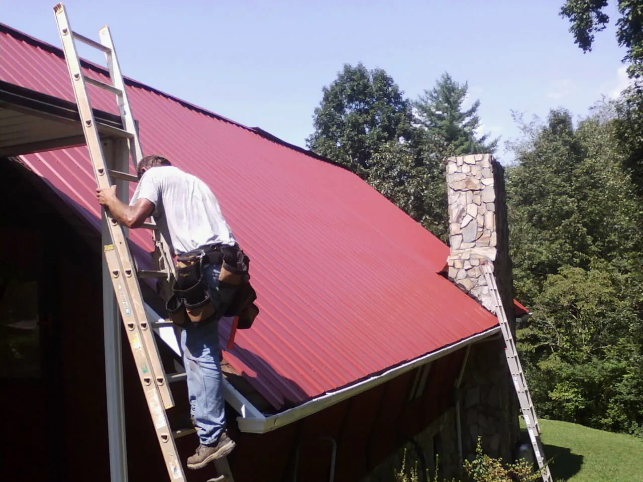 Roofer on a ladder beside a red metal roof and stone chimney — Soffit & Fascia Repair by Roofing Brothers in Nordland